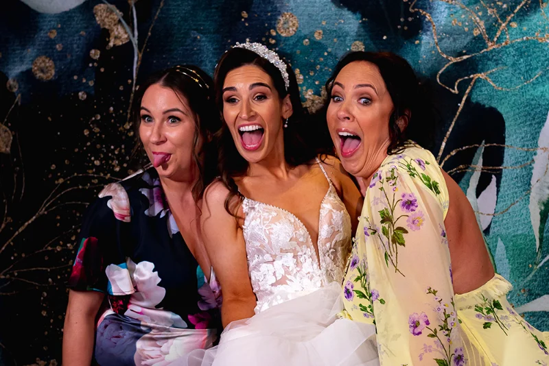 Une mariée et deux invitées rient devant un photobooth lors d'un mariage